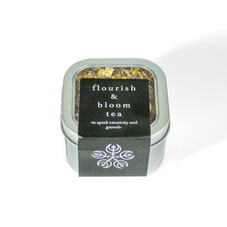 Flourish and Bloom tea in square tin container with see through front, showing loose dried flowers. Black label with sigil. 