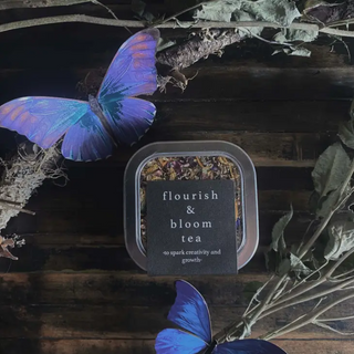 Flourish and Bloom Tea in square tin container with black label. Shown on wooden background with purple butterflies and dried leaves.