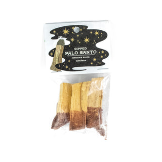 Clear package holding three sticks of palo santo, the bottom half of each is red and covered in loose herbs. The label at the top shows a drawing of a lit palo santo stick with a black smoke swirl with stars in it and says Dipped Palo Santo