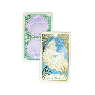 The Moon card from the Ethereal Visions Tarot deck shows the outline of a woman in white and gold against a starry blue sky. A second card is behind it showing the back side of the cards. There are purple spheres with green ferns. 