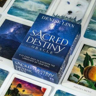 Box of the Sacred Destiny Oracle is blue and shows a volcano erupting in a night sky. Other cards from the deck are surrounding it.