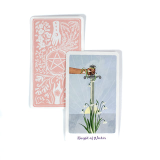 The Knight of Winters card from the Harmony Tarot deck shows a hand holding a sword pointed into the ground into flowers. A second card shows the back of the deck, it is light pink with white drawings of two hands reaching toward a pentagram with herbs and flowers around it. 
