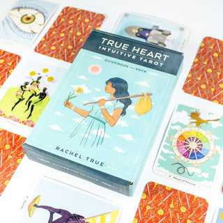 True Heart Intuitive tarot box is light blue and show a woman with dark hair holding a flower and a knapsack over her shoulder. Other cards from the deck are shown surrounding the box.