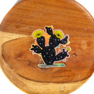 Sticker of a the outline of a prickly pear cactus filled in with black and stars showing the universe inside. Cactus flowers are yellow with eyes. Sticker is holographic and shown on wooden plate.