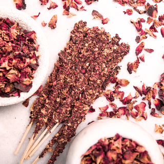 Several extra thick rose covered incense sticks. There are small bowls filled with rose petals next to it and rose petals scattered in the background.