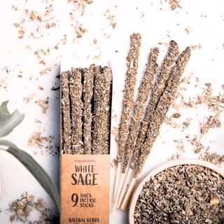 White Sage thick floral and herb incense sticks shown in packaging, with several sticks ourside the package. There is also a bowl full of loose herbs on a white background.