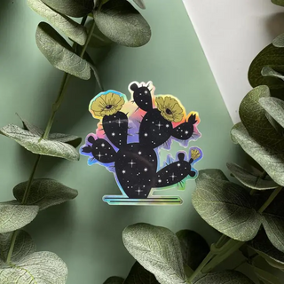 Sticker of a the outline of a prickly pear cactus filled in with black and stars showing the universe inside. Cactus flowers are yellow with eyes. Sticker is holographic and shown a green background with leaves.