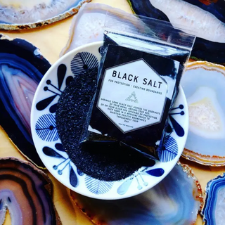 Small plastic baggie filled with black salt. Label is black and white with uses- for protection and creating boundaries. Bag is in a bowl filled with the black salt with agate slabs as the background.