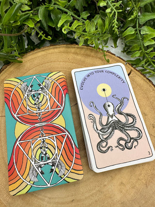 Two stacks of Animal Apothecary Oracle cards. One shows the back of the cards, which has a turquoise background and red and orange rainbows. There are mirrored images of a Caduceus symbol. The front image of the card shows a black and white octopus drawing on light pastels with the text Evolve Into Your Complexity.