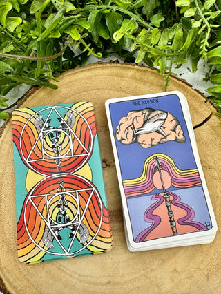 Two stacks of Animal Apothecary Oracle cards. One shows the back of the cards, which has a turquoise background and red and orange rainbows. There are mirrored images of a Caduceus symbol. The front image of the card shows a a drawing of a shark inside of a brain with a spear pointing towards it with the text The Illusion.