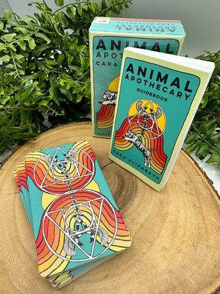 The Animal Apothecary Oracle card box with guidebook shown. There is a stack of cards showing the back of the cards on a wooden background. 