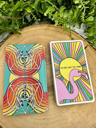 Two stacks of Animal Apothecary Oracle cards. One shows the back of the cards, which has a turquoise background and red and orange rainbows. There are mirrored images of a Caduceus symbol. The front image of the card shows a drawing of a pink flamingo with different colored rays coming out of a sun behind it with the text You Are What You Think.