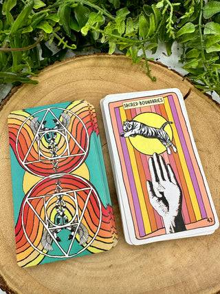 Two stacks of Animal Apothecary Oracle cards. One shows the back of the cards, which has a turquoise background and red and orange rainbows. There are mirrored images of a Caduceus symbol. The front image of the card shows a drawing of a black and white hand with a tiger jumping above it with the text Sacred Boundaries.
