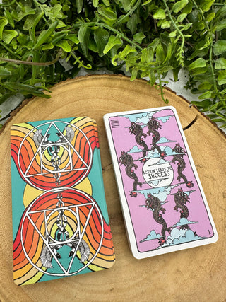 Two stacks of Animal Apothecary Oracle cards. One shows the back of the cards, which has a turquoise background and red and orange rainbows. There are mirrored images of a Caduceus symbol. The front image of the card shows several seahorses on a lavender background with the text "Action Leads to Success."