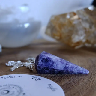 Purple lepidolite pendulum laying on its side next to a pendulum grid and crystals.