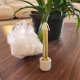 Gold metallic chime candle in white chime holder next to large chunk of raw clear quartz and a plant.