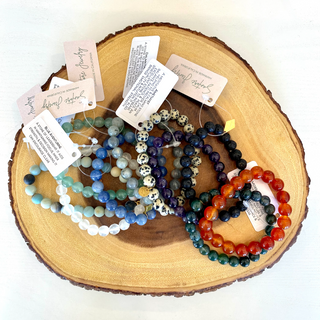 Ten varieties of crystal mala gemstone bracelets placed together on a wooden plate. Each has a tag indicating the crystals metaphysical properties. 
