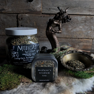 Tin of Medusas Wisdom herbal tea with a large jar of loose leaf tea next to it, and a brass bowl with loose herbs inside. There is a brass statue of Medusa behind them. 