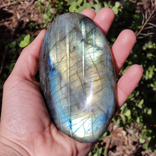 Hand holding a large labradorite palm stone, light blues and greens with flashes of orange.